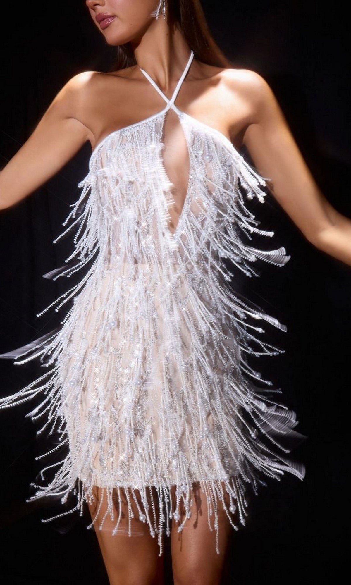 A person models the Ladivine CDS574, a short off-white homecoming dress with a halter neckline, fringe beadwork, and keyhole cutout, standing against a dark background.